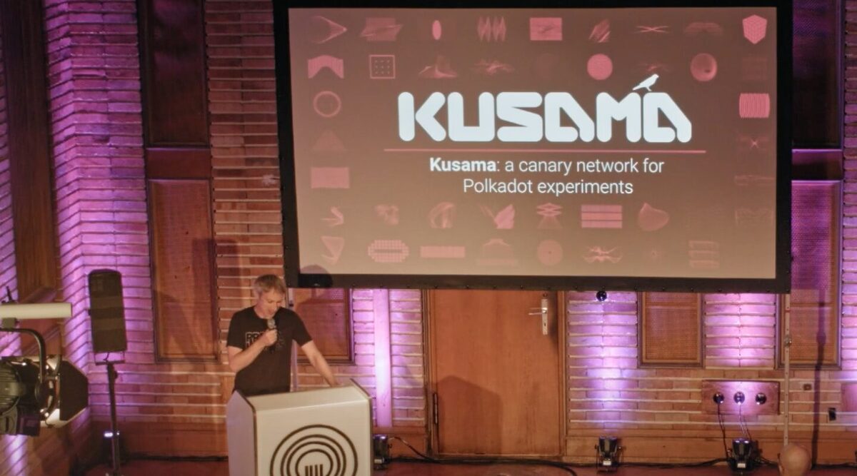 Gavin Wood, the founder of Ethereum, standing in a conference with a board behind him that says "Kusama: A canary network for Polkadot experiments"