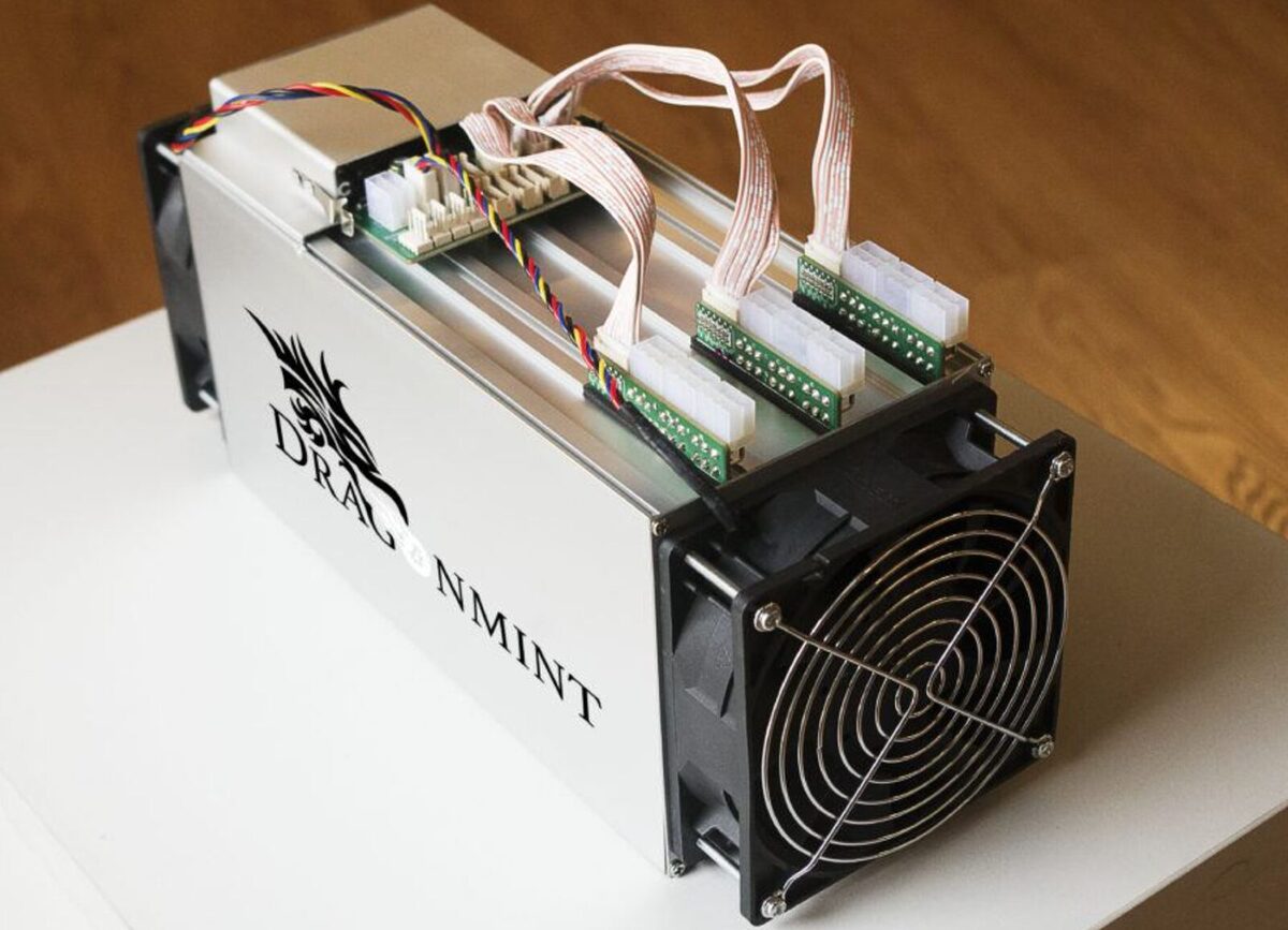Silver Dragonmint branded ASIC rig which is exposed at the top to show cables and wires coming out of it. The rig is resting on a small silver table.