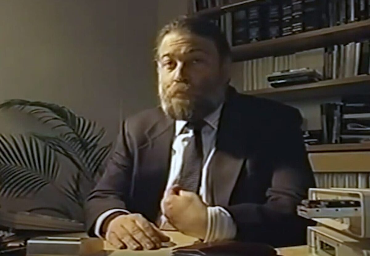 David Chaum in a business suit sat next to a desk. He is surrounded by books and documents in an office environment.