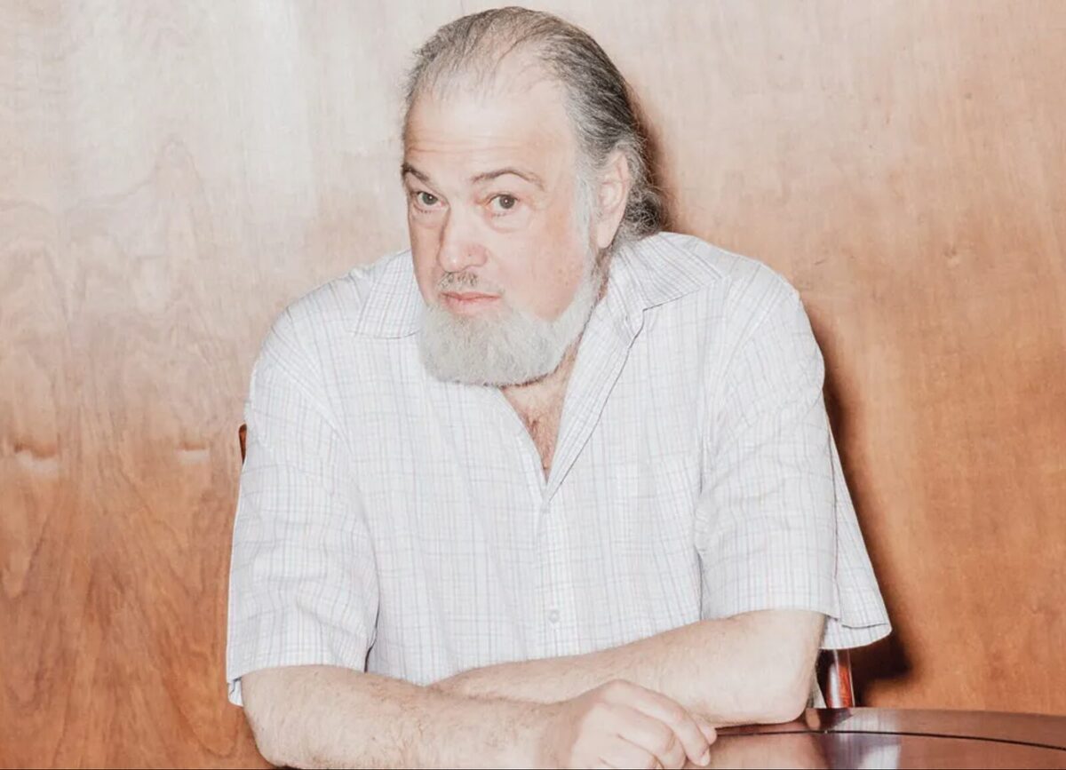 David Chaum sitting with his arms folded on a desk. He is wearing a white shirt and has a suprrised look on his face.