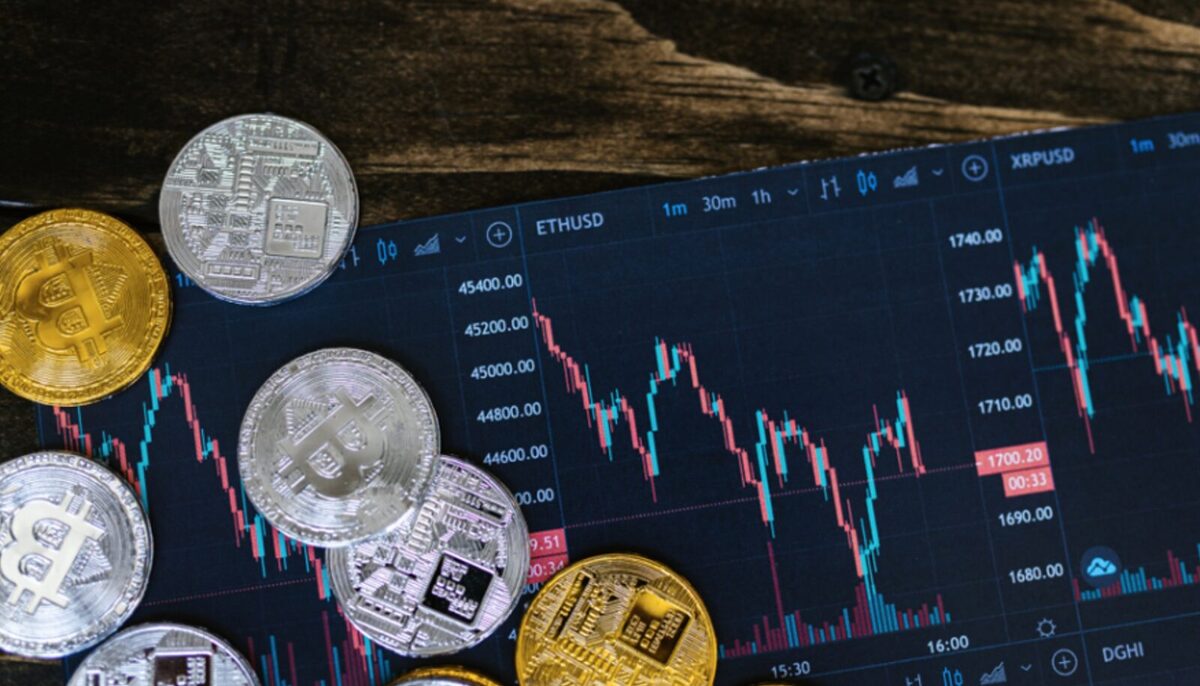 Multiple silver and gold Bitcoins on the left and a market graph showing its value going up and down in the background.