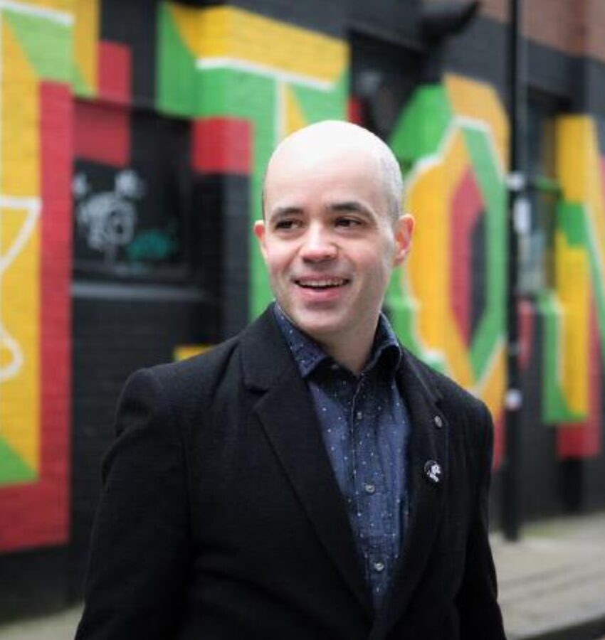 Ross Nicoll laughing at something offscreen. He is wearing a black blazer and blue undershirt and standing in front of a colorful street wall.