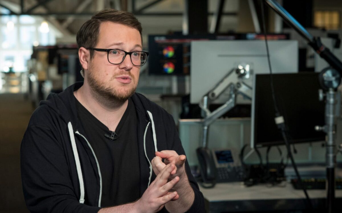 Jackson Palmer speaking to an interviewer who is off-camera. He is in an office with computers in the background.