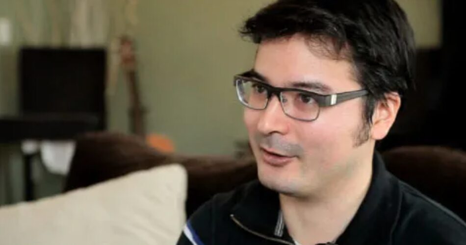 Billy Markus speaking to an interviewer who is off-screen. He is wearing a black polo shirt and black glasses.