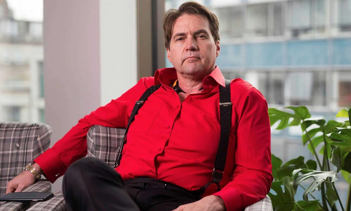 Craig Wright in a red shirt sitting on a chair with a smirk on his face.