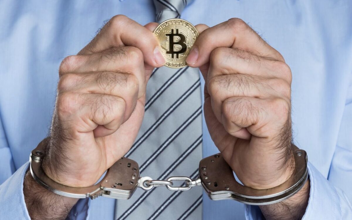 A handcuffed pair of hands holding up a golden Bitcoin token.