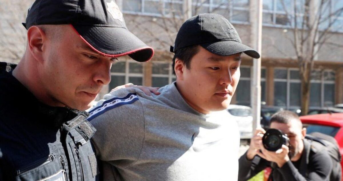 Do Kwon with a grey jumper and baseball cap being escorted from a building by a policeman. A man with a camera can be seen in the background taking photos of the arrest.