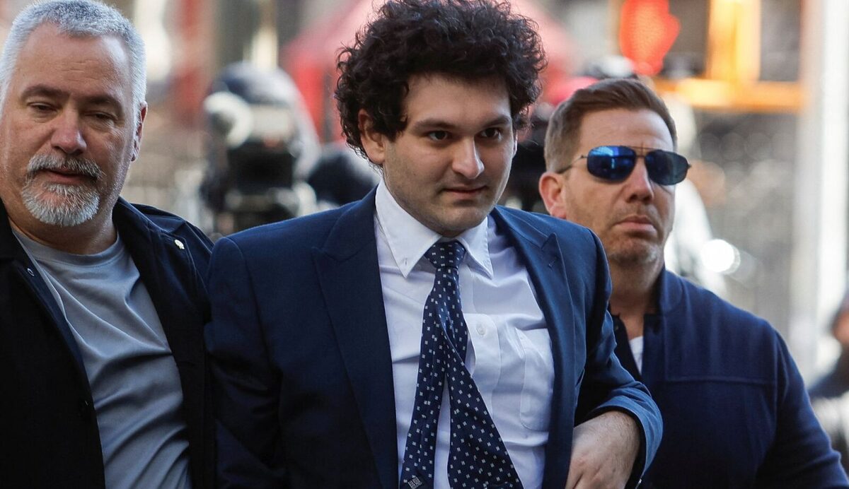 Sam Bankman-Fried being escorted by two smart looking lawyers. Bankman-Fried is wearing a blue blazer and dotted tie.