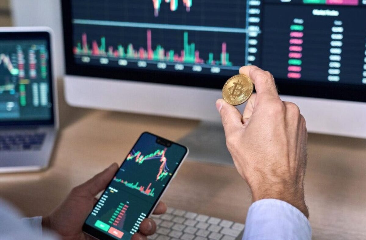 Man with a phone showing a decentralised exchange market with a decentralised exchange also on a desktop in the background. One hand is holding a gold Bitcoin token.
