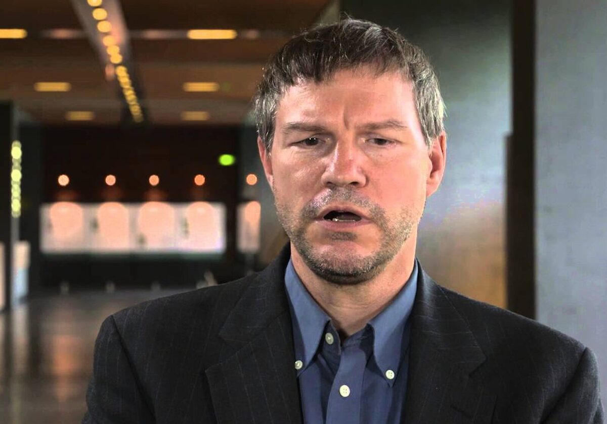 Nick Szabo in a black blazer and blue undershirt speaking on camera in an empty studio.