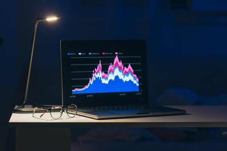 Computer in a dark room placed on a table next to a turned on desk lamp and glasses.