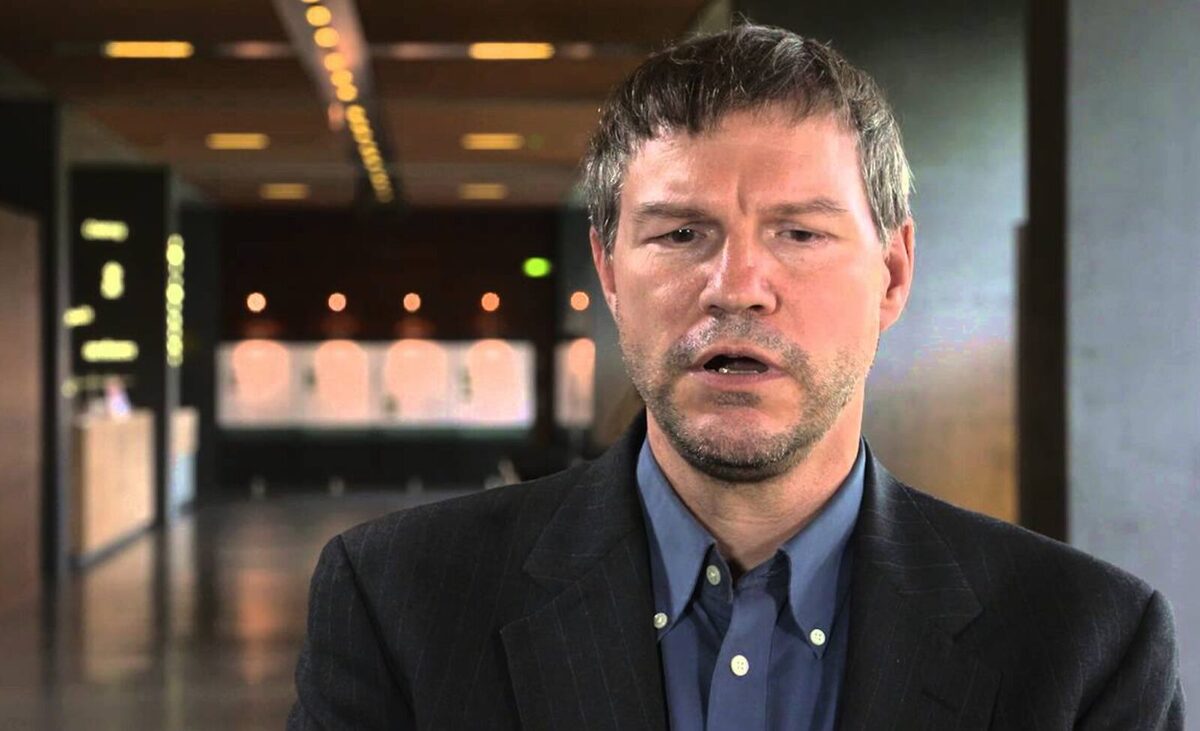 Nick Szabo in a black suit and blue undershirt speaking to the camera while standing in an empty hallway.